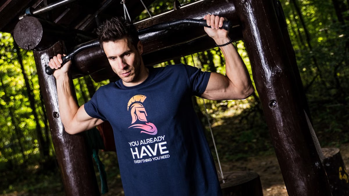 A man in a motivational t-shirt using an outdoor gym in a forest setting.